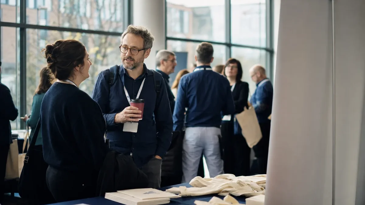 People with lanyards talking in a bright conference foyer near a table of printed programmes