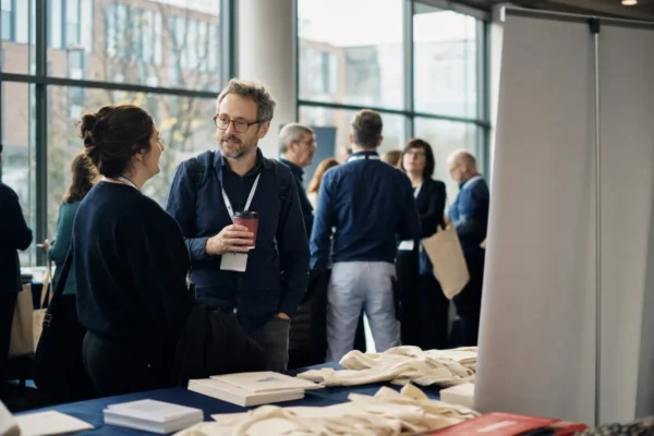 People with lanyards talking in a bright conference foyer near a table of printed programmes
