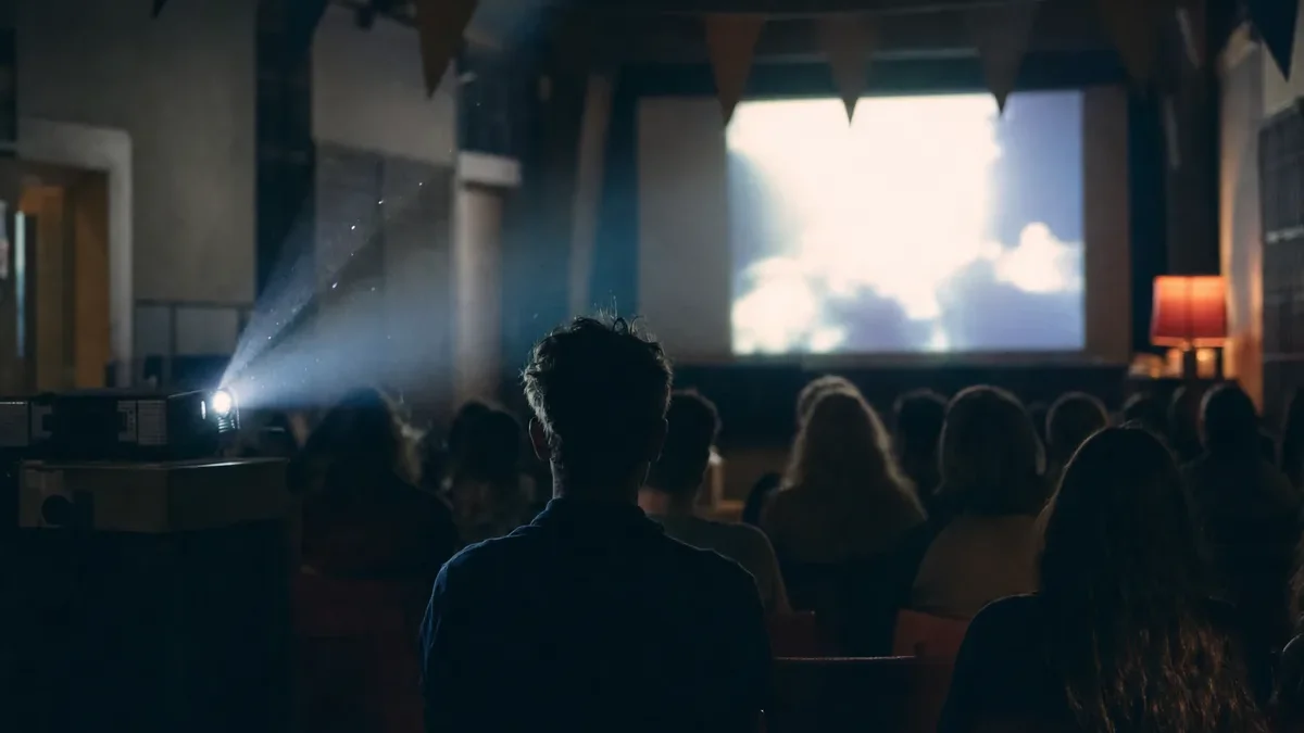 Audience in a dim hall watching a bright projection screen with a projector beam overhead