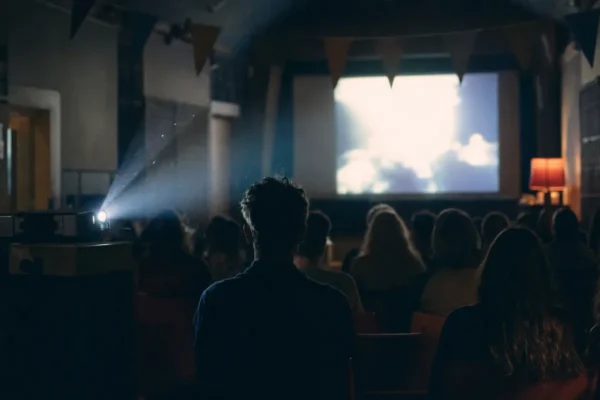 Audience in a dim hall watching a bright projection screen with a projector beam overhead